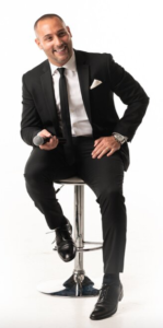 A man in black suit and tie sitting on a stool.